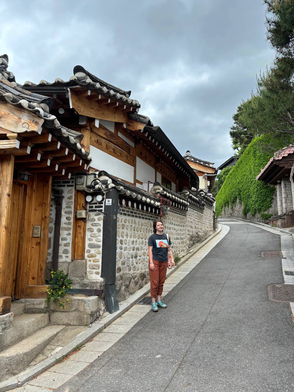 Veronika standing on the side of a small street near a wood and stone building in South Korea, wearing a gray shirt and dark orange pants