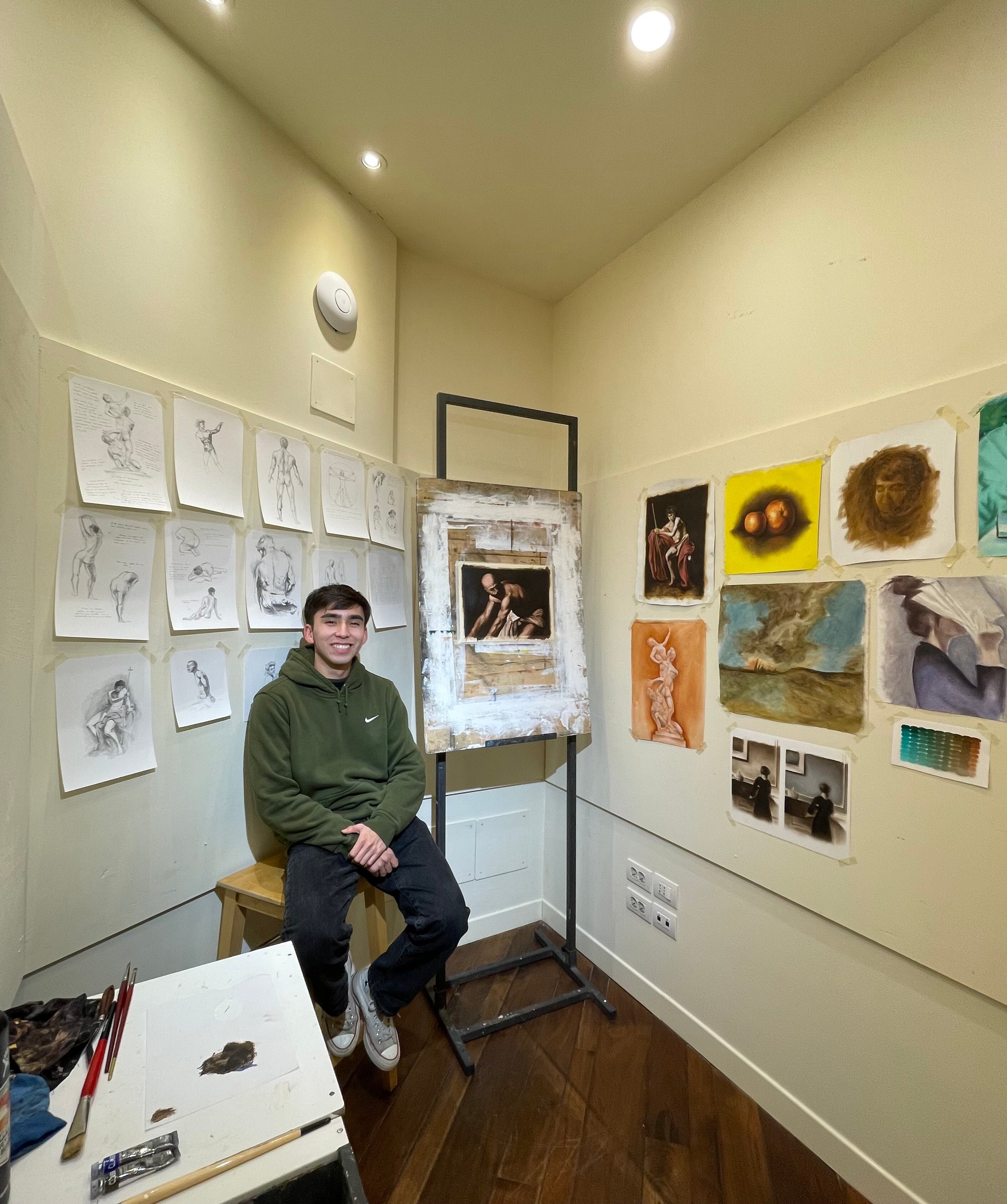 Yael sits in a green hoodie on a stool next to his painting in a small studio space with various other pieces of art on the walls.